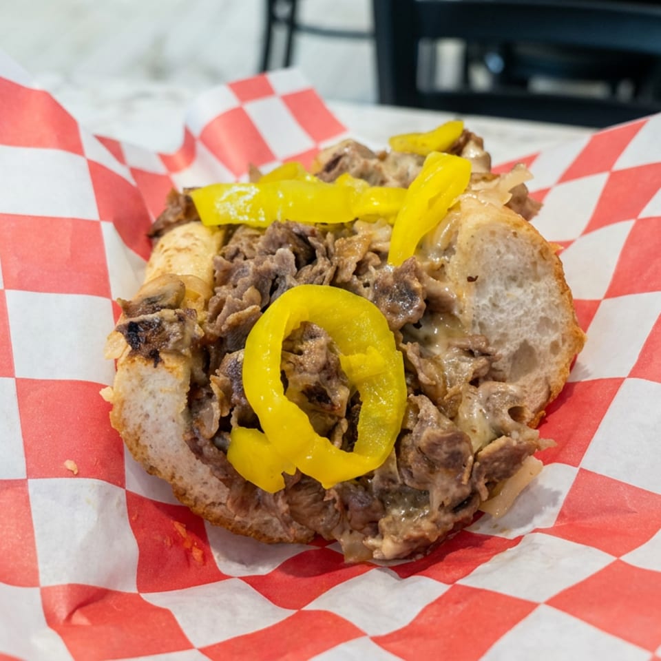 Cheesesteak sandwich piled with chopped beef, melted cheese, mushrooms, and yellow peppers on red-and-white checkered paper.