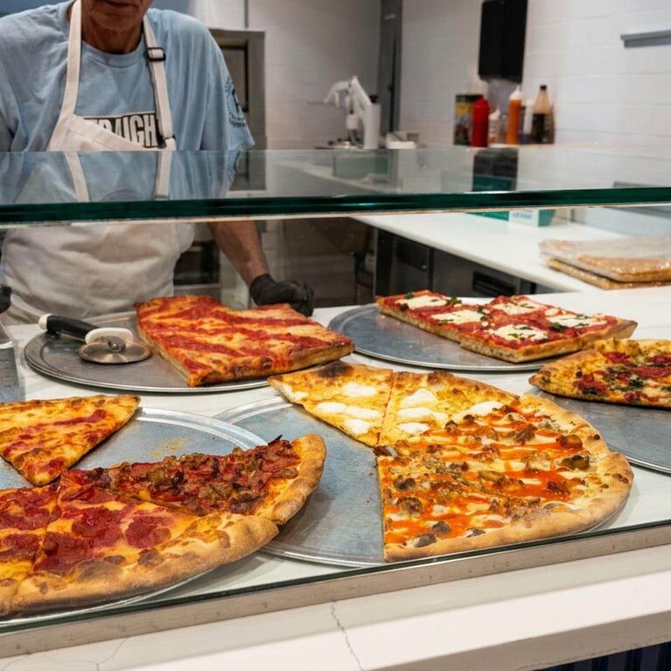 Restaurant worker serves assorted rectangular and round pizzas behind a glass display counter.