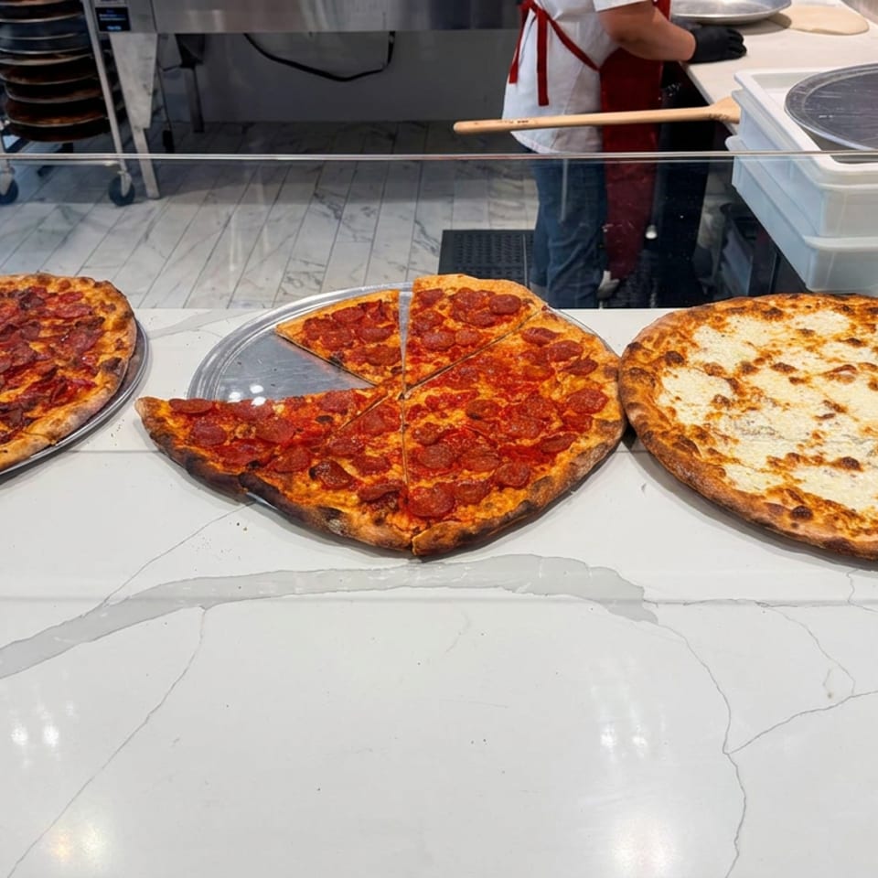 Three pizzas sit on metal trays at a restaurant counter: two topped with pepperoni and one plain, with a worker visible behind the counter.