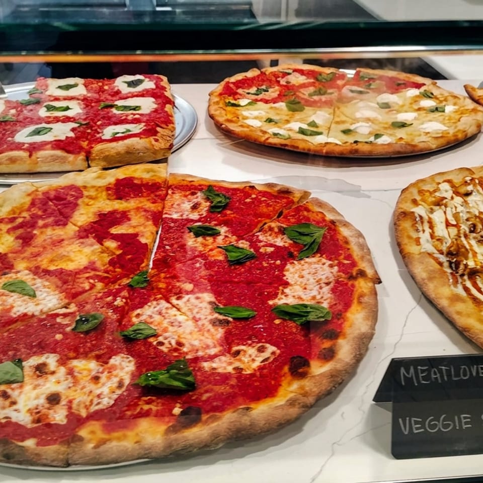 Assorted pizzas, including Margherita and specialty varieties, displayed in a restaurant glass counter with handwritten slice labels.